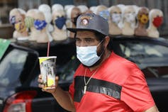 Un homme en chemise rouge, masqué, tient un soda devant un étalage de mannequins portant tous des masques