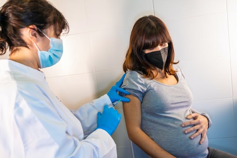 femme enceinte masquée se faisant vacciner par une femme en blouse blanche, masquée elle aussi.